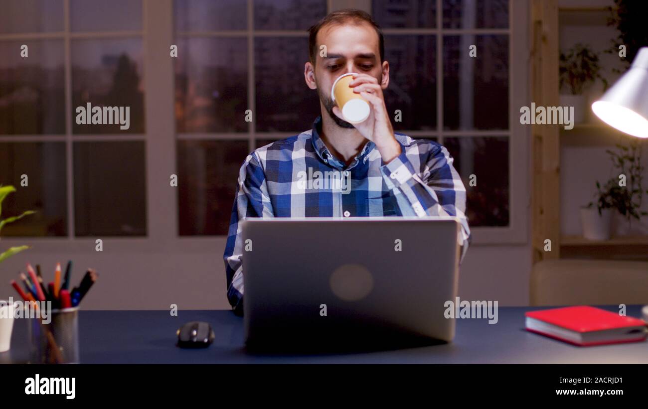 Imprenditore barbuto prendendo un sorso di caffè e di lavoro sul computer nel suo ufficio di casa durante le ore notturne. Foto Stock