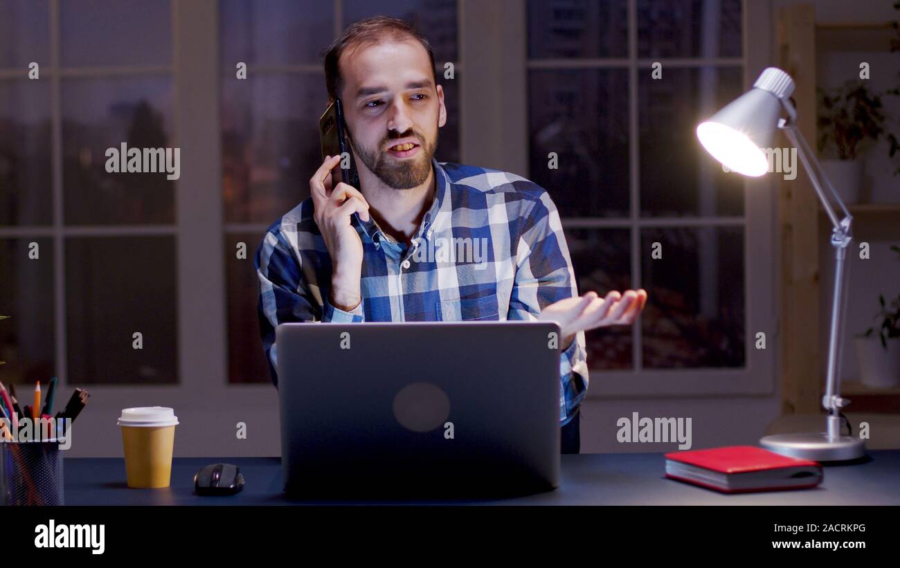 Imprenditore avente un argomento di conversazione al telefono mentre si lavora nel suo ufficio di casa durante le ore notturne. Foto Stock
