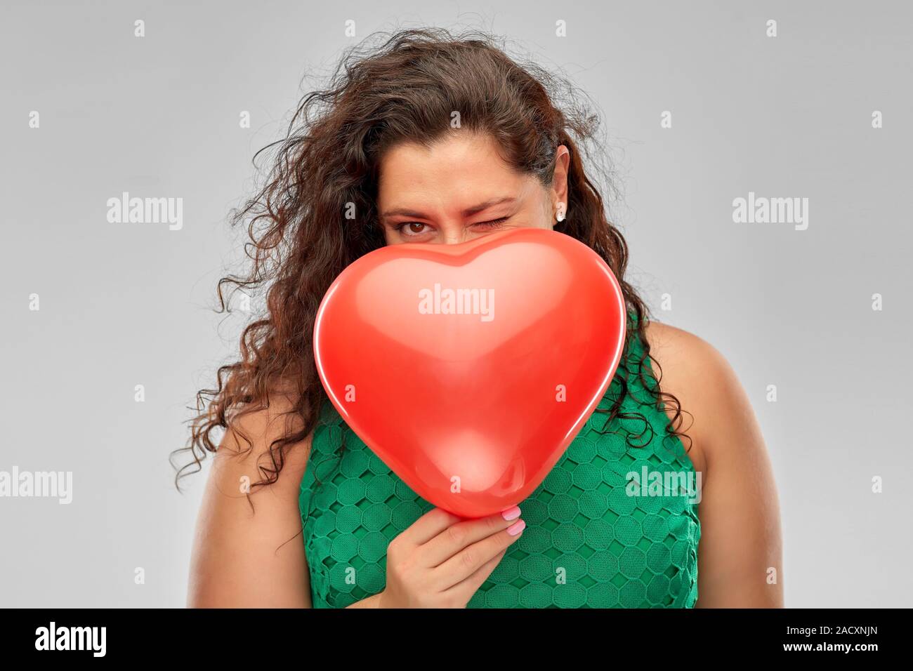 Il giorno di san valentino, le persone e il concetto di amore - giocosa donna in abito verde di nascondersi dietro a cuore rosso a forma di palloncino su sfondo grigio Foto Stock