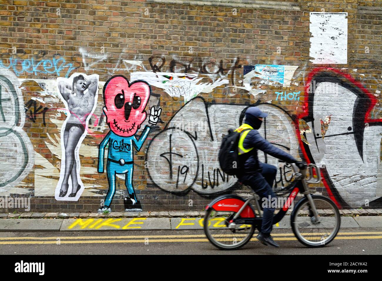 Un ciclista passando graffiti su un muro di mattoni in Buxton Street off Brick Lane Whitechapel Londra Inghilterra REGNO UNITO Foto Stock