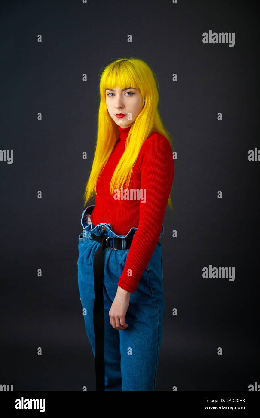 Una giovane donna con colore giallo brillante capelli indossando un rosso brillante a manica lunga in cima contro un semplice sfondo scuro. Foto Stock