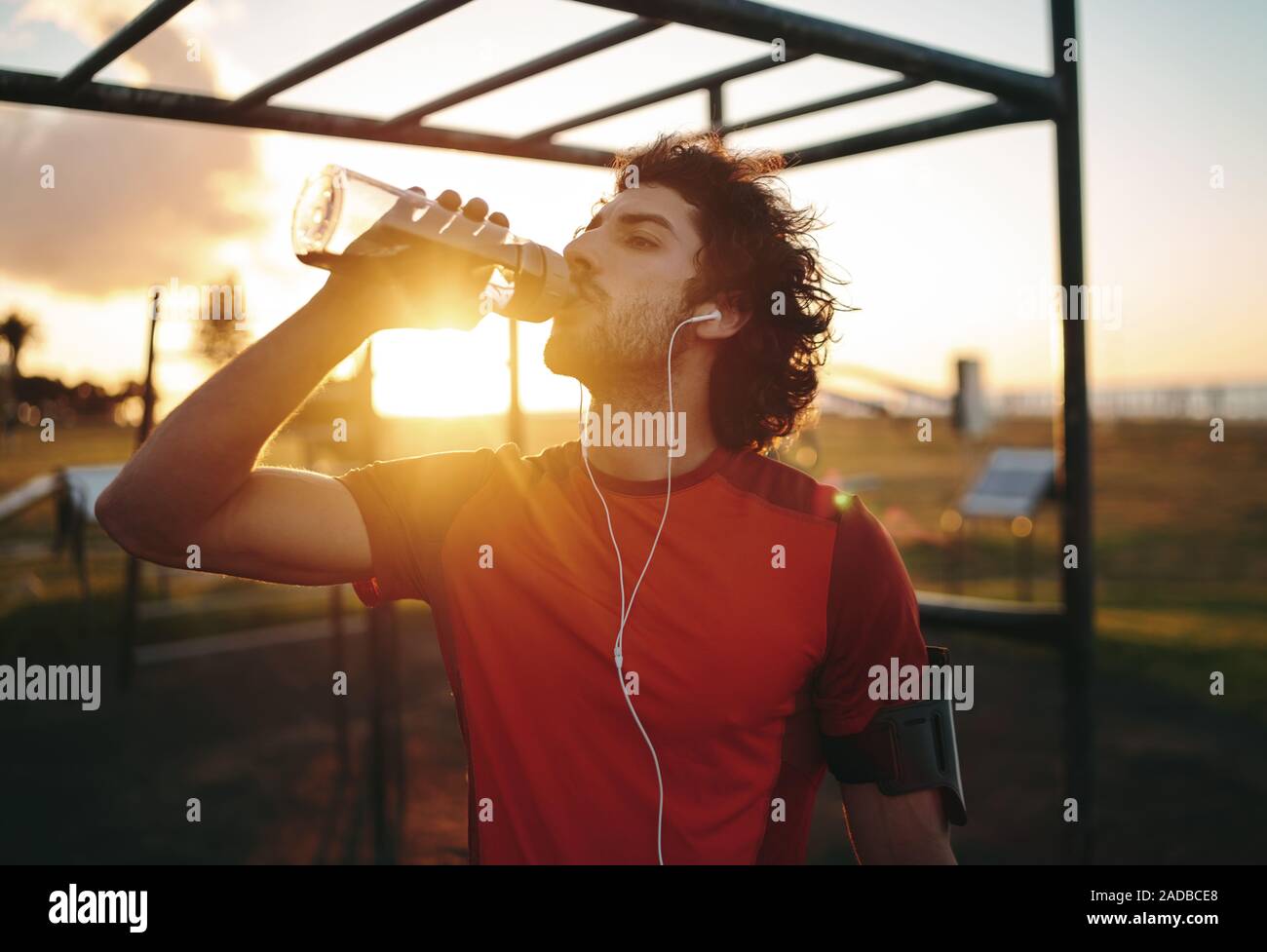 Fitness atleta giovane uomo con gli auricolari nelle orecchie di bere acqua da una bottiglia riutilizzabile dopo allenamento esercitando su estate in palestra park Foto Stock