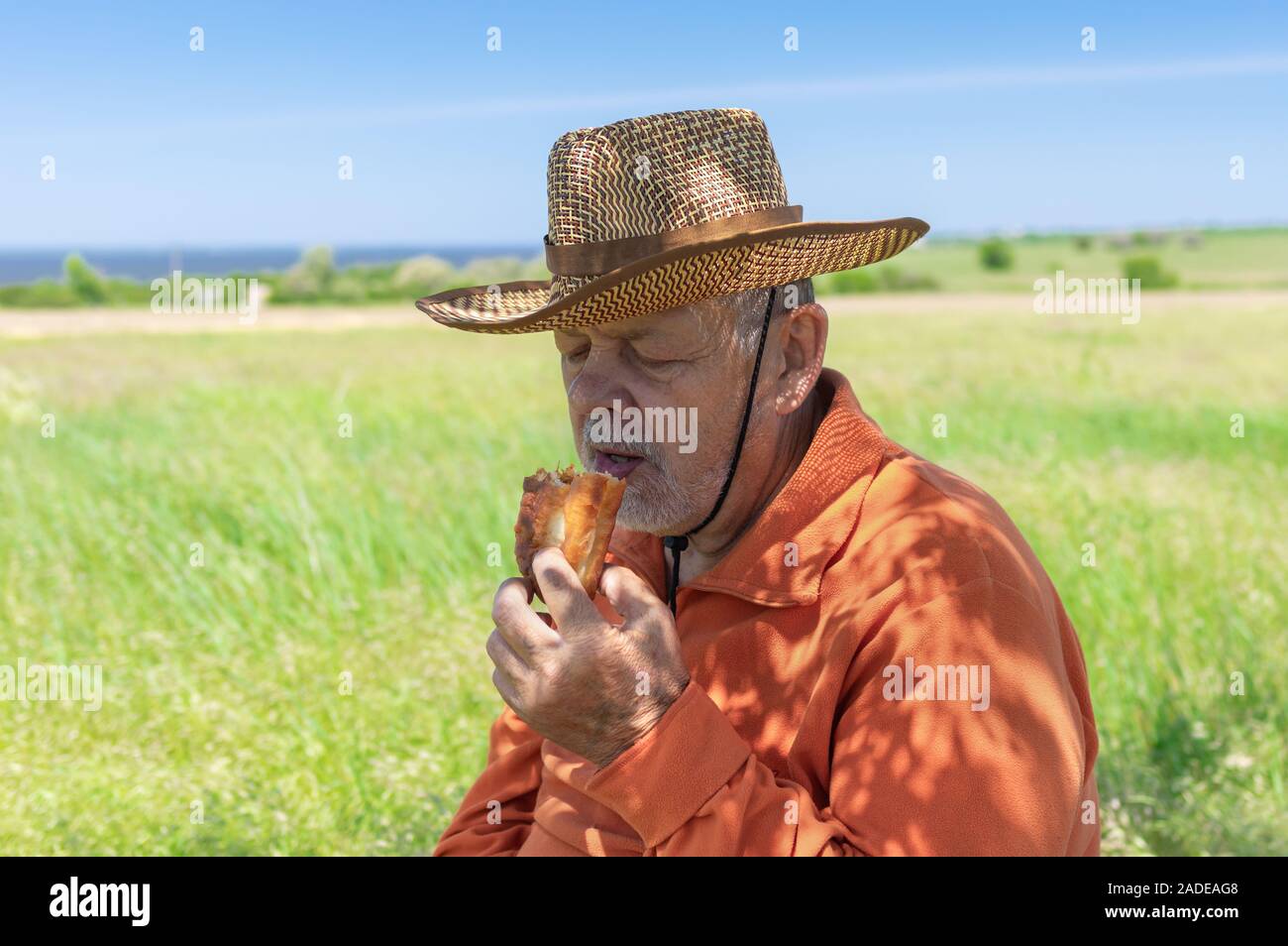 Fame caucasica agricoltore senior mangiare artigianale di Patty sotto ombra di albero vicino al suo campo Foto Stock