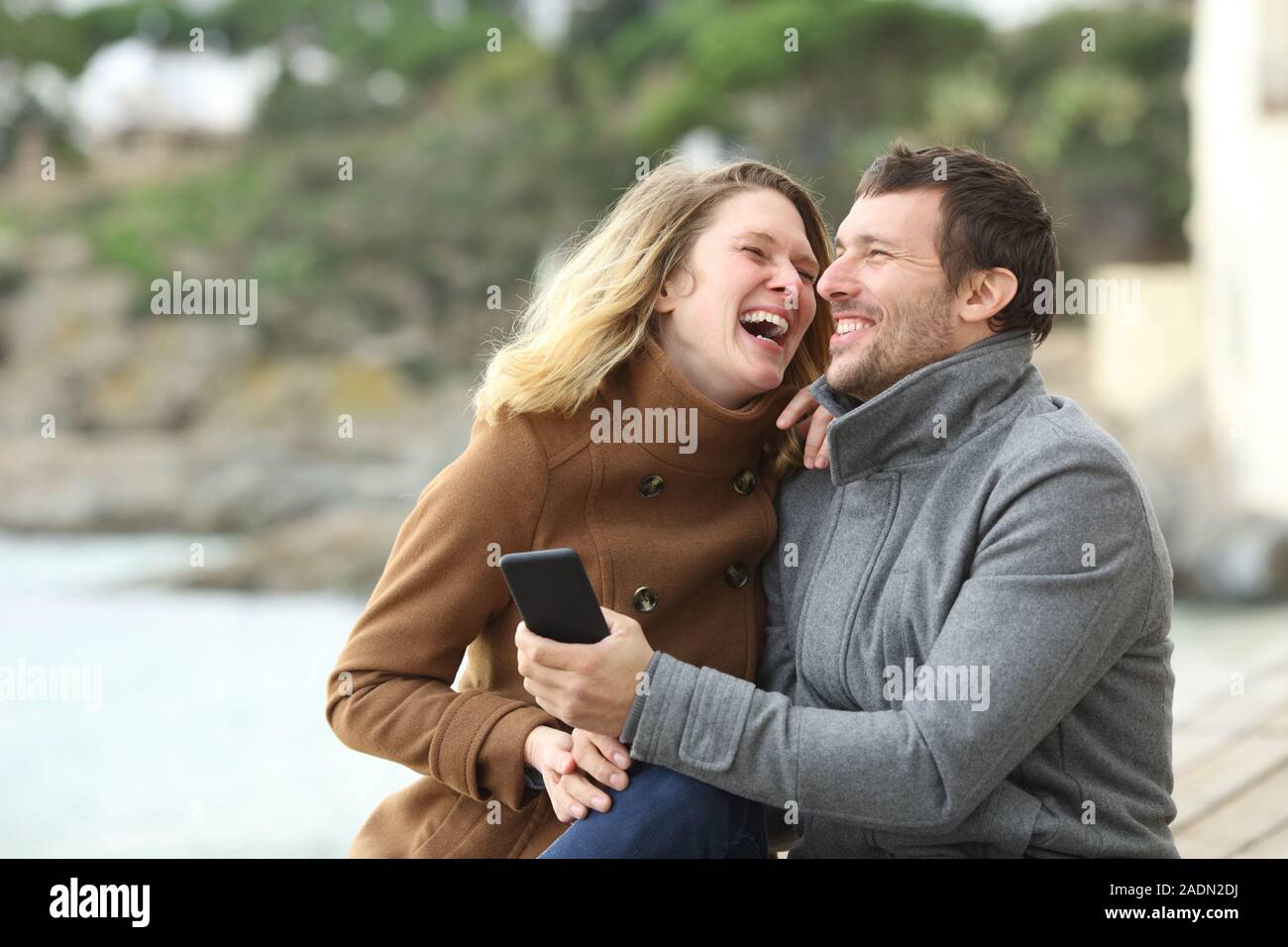 Coppia felice di adulti ridevano forte su telefono cellulare contenuto seduto sulla spiaggia in inverno Foto Stock