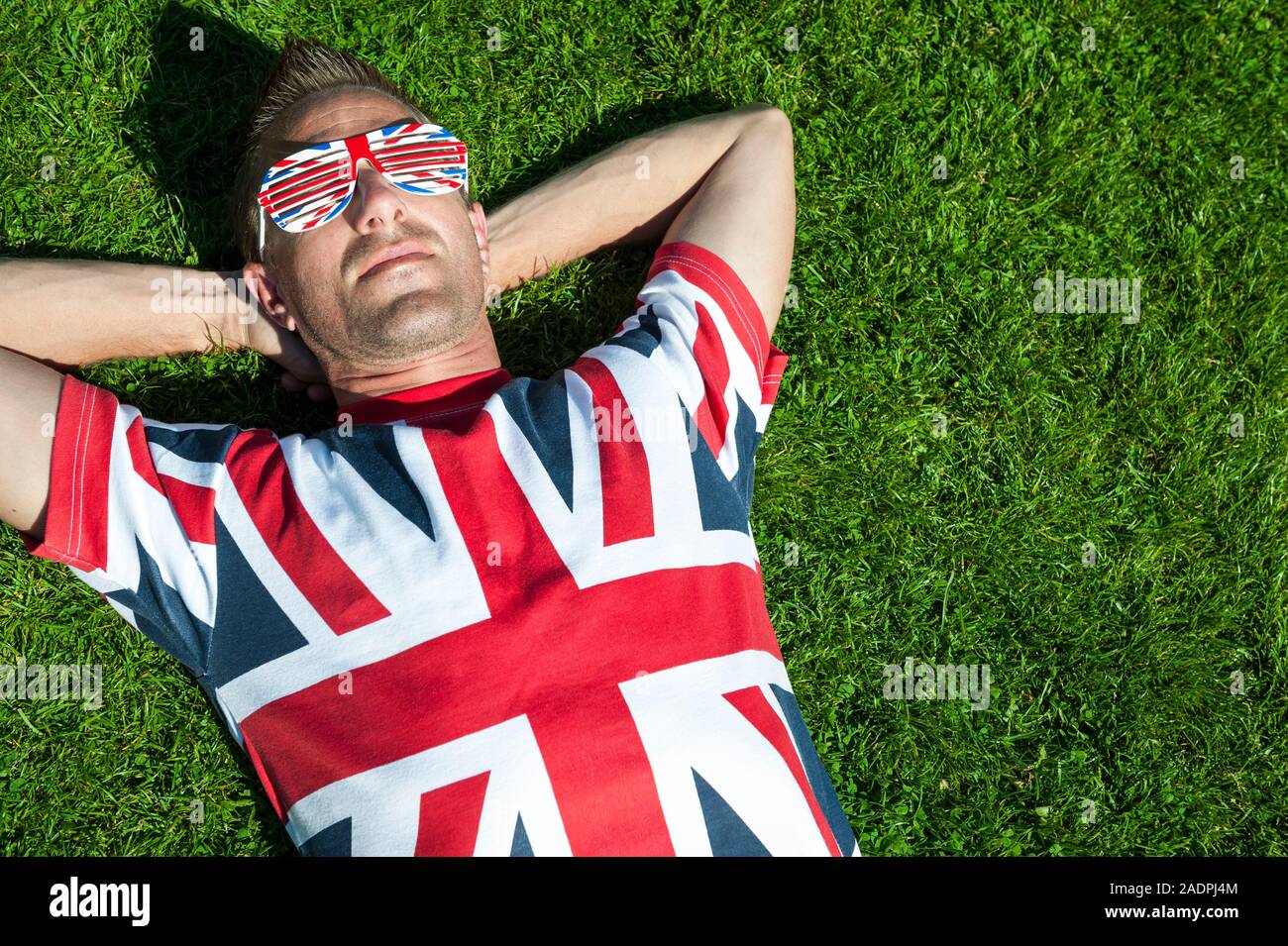Rilassata giovane britannico in Unione Jack T-shirt e occhiali da sole si trova indietro su un soleggiato patch di estate verde erba Foto Stock