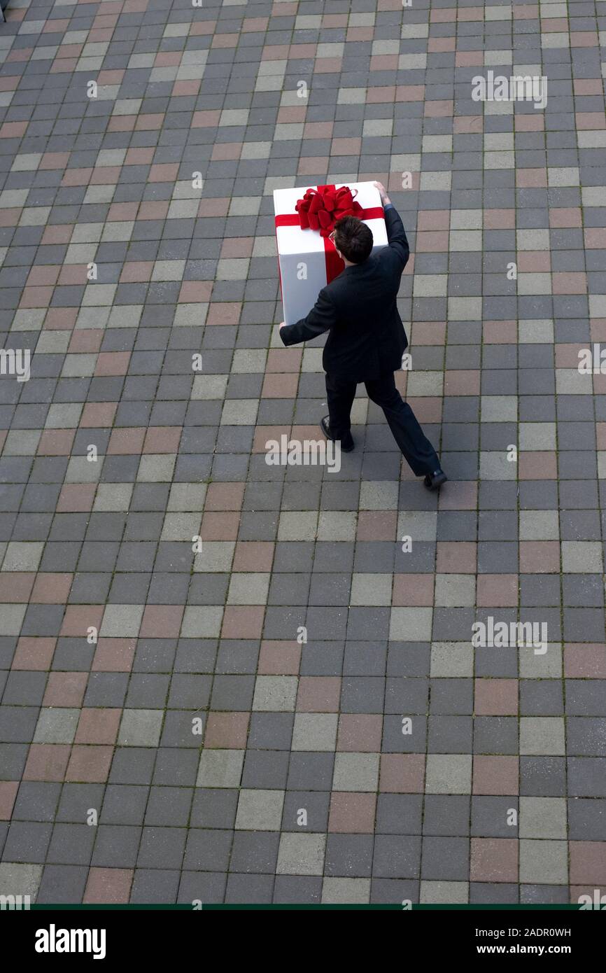 Un giovane uomo che porta un grande regalo bianco avvolto con un fiocco rosso passeggiando attraverso un cortile Foto Stock