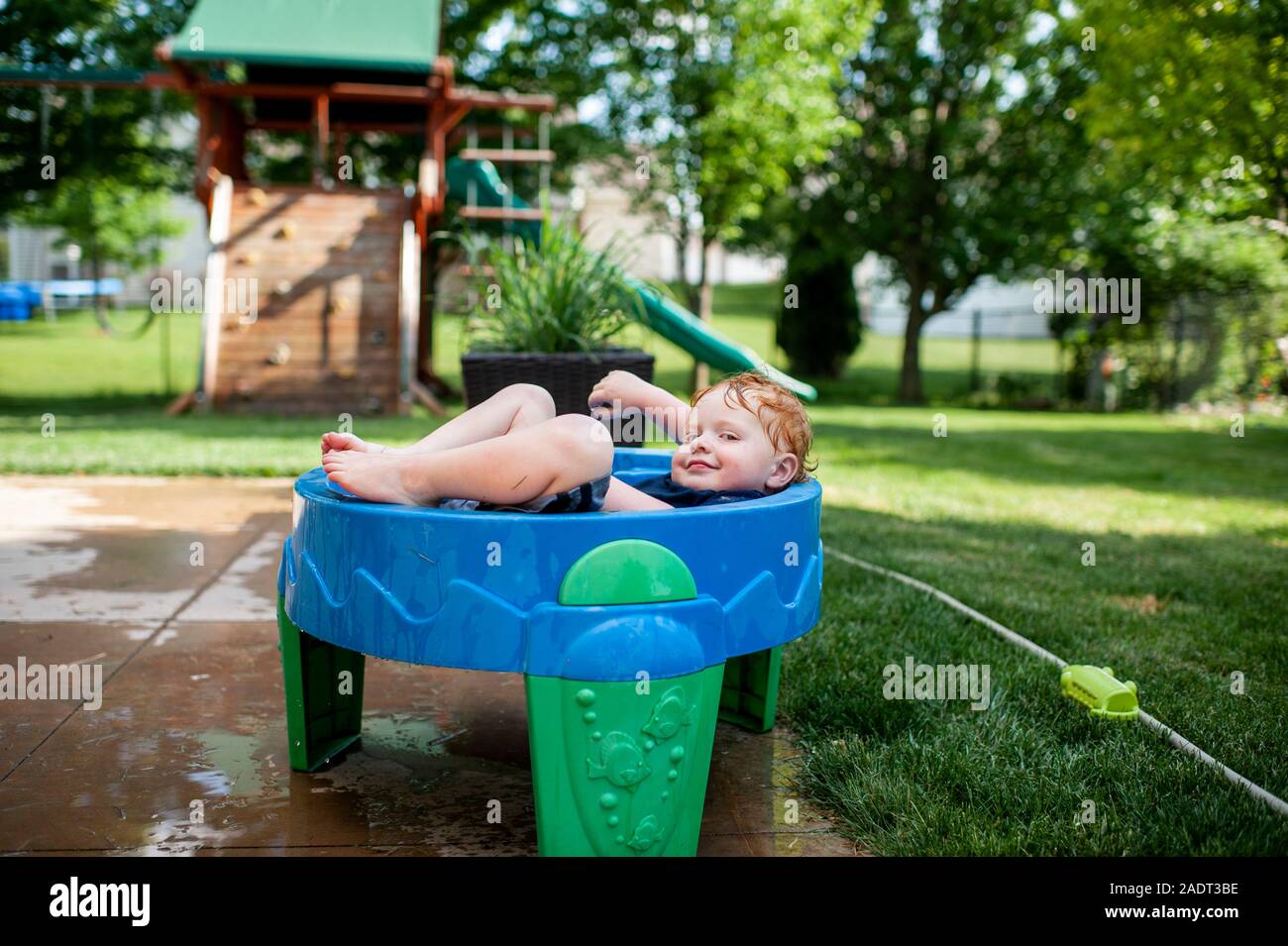 Il Toddler boy smirking come egli getta in acqua a tavola con vestiti all'esterno Foto Stock