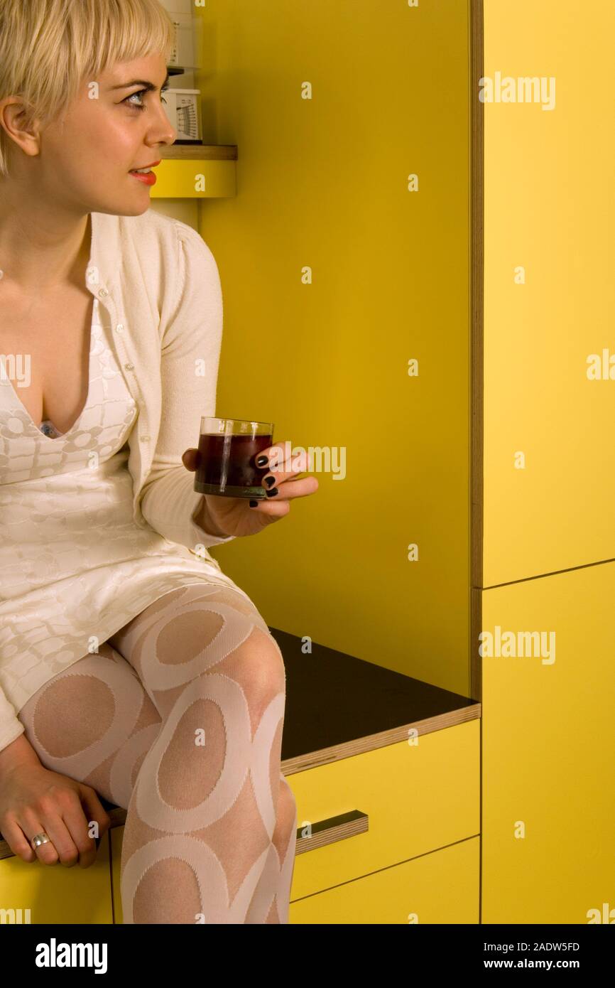 Una giovane donna, 20's, seduto su un piano di lavoro di una cucina con in mano un bicchiere di vino o di succo di frutta e interagire con un'altra persona al di fuori della fotocamera Foto Stock