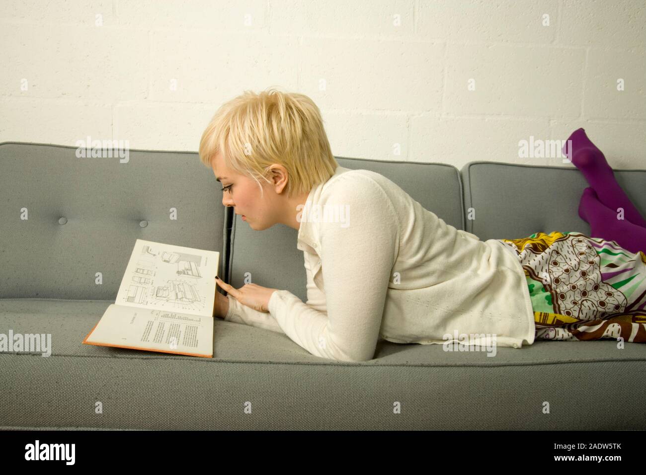 Elegante giovane donna, 20's, in un semplice degli anni sessanta stile retrò interni domestici, la lettura di un libro, sdraiato su un divano Foto Stock