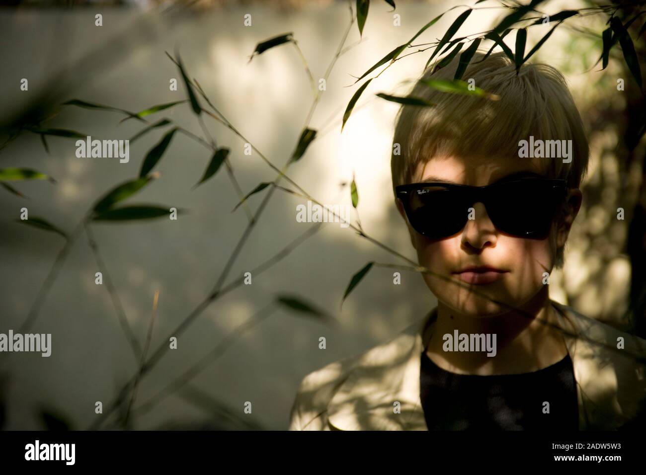 Elegante giovane donna, 20's, indossando stile anni '50 trench e occhiali da sole, visto attraverso fogliame Foto Stock