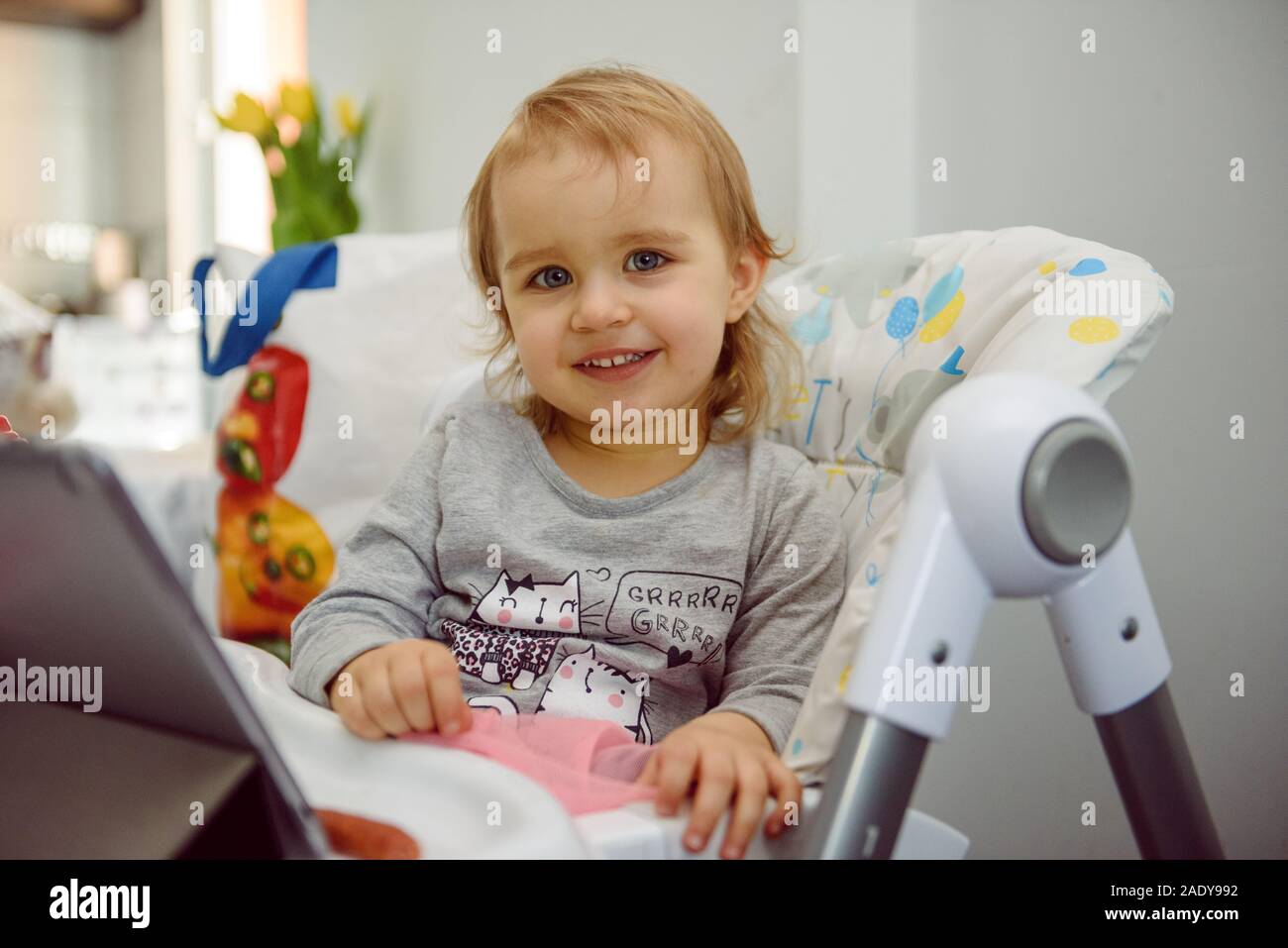 Carino bambino biondo si siede in un seggiolino per bambino in cucina a mangiare una succosa pesca. Bambina con un piercing look. Foto Stock