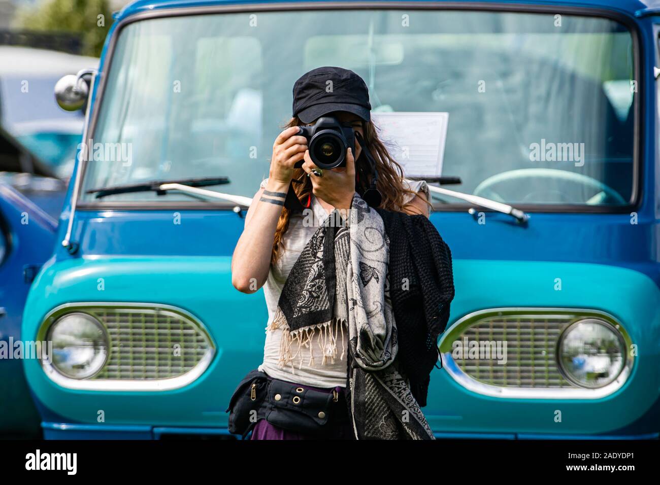 Messa a fuoco selettiva su giovani caucasici donna fotografo tiene la fotocamera reflex digitale nella parte anteriore del vecchio blu van, indossare un cappello nero sciarpa, durante un'auto d'epoca mostrano Foto Stock