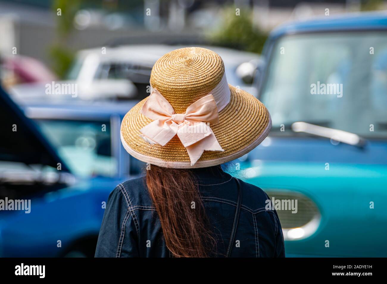 Vista posteriore della donna che indossa una giacca di jeans e cappello per il sole con larga tesa come si guarda a autovetture durante la outdoor antique car show, offuscata sfondo auto Foto Stock