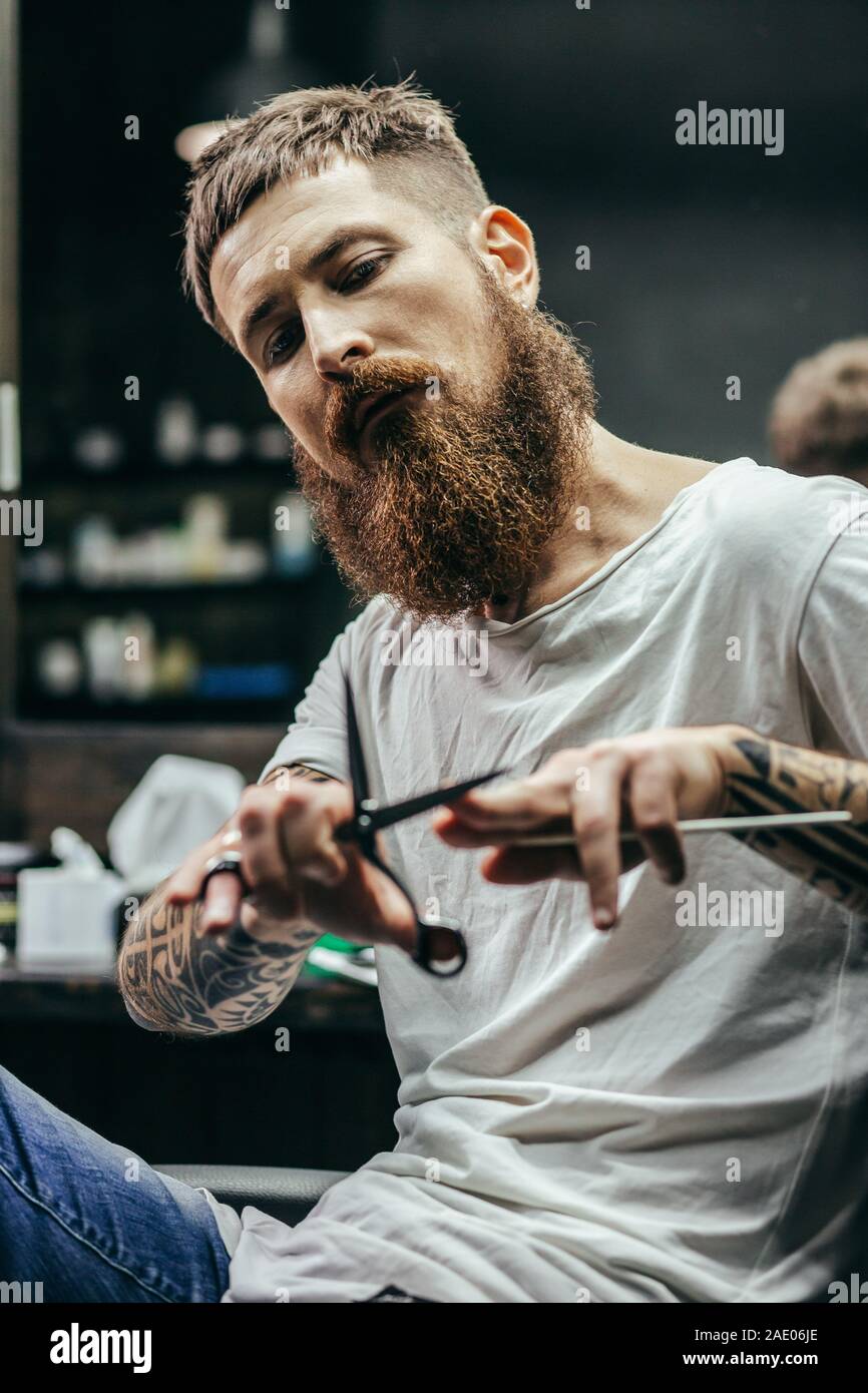 Barbiere pensieroso seduto e toccando il suo forbici Foto Stock