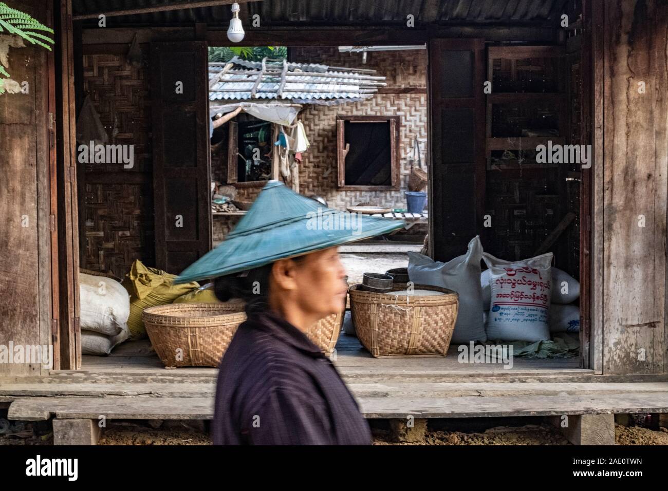 Una donna più anziana in un riso conica hat passa da un magazzino rurale in un composto su un villaggio street nelle zone rurali del nord-ovest del Myanmar (Birmania) Foto Stock