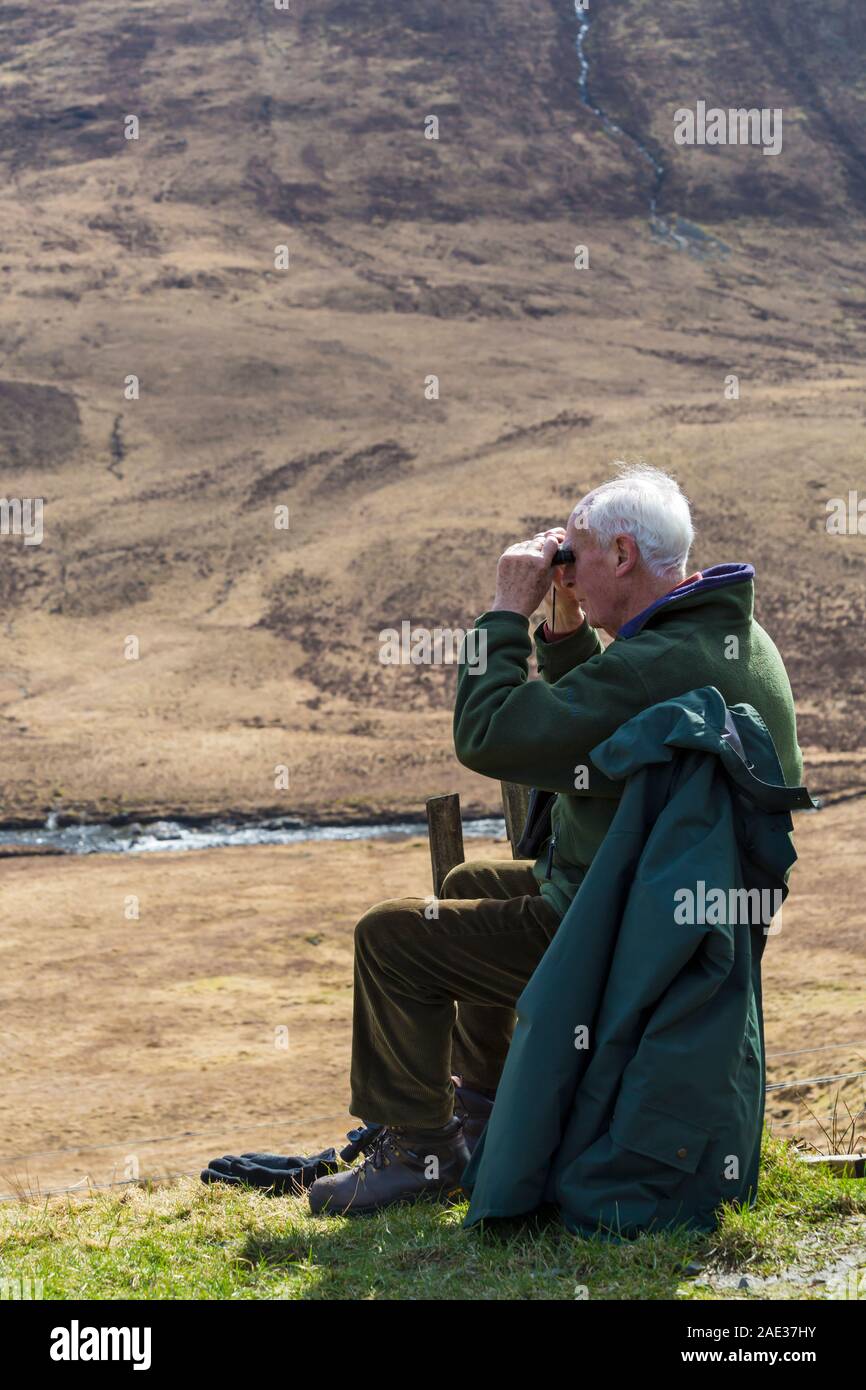 Uomo seduto su un banco di lavoro guardando attraverso il binocolo alla Fairy Piscine, Isola di Skye in Scozia, nel Regno Unito nel mese di marzo Foto Stock