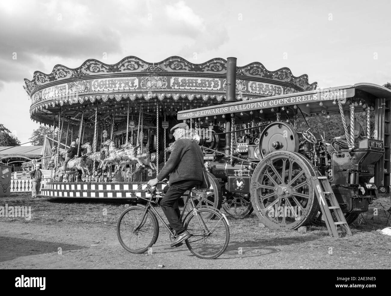 Il vecchio uomo in bicicletta pedala in bicicletta attraverso una fiera tradizionale a vapore d'epoca con giostra e motore di trazione Malpas Inghilterra Foto Stock