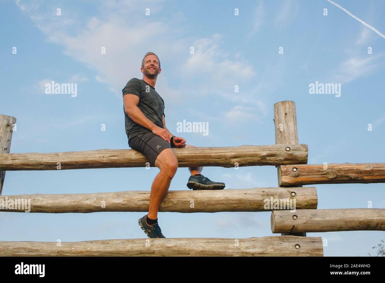 Un orgoglioso uomo sorridente siede sulla cima di un alto recinto in legno contro il cielo blu Foto Stock