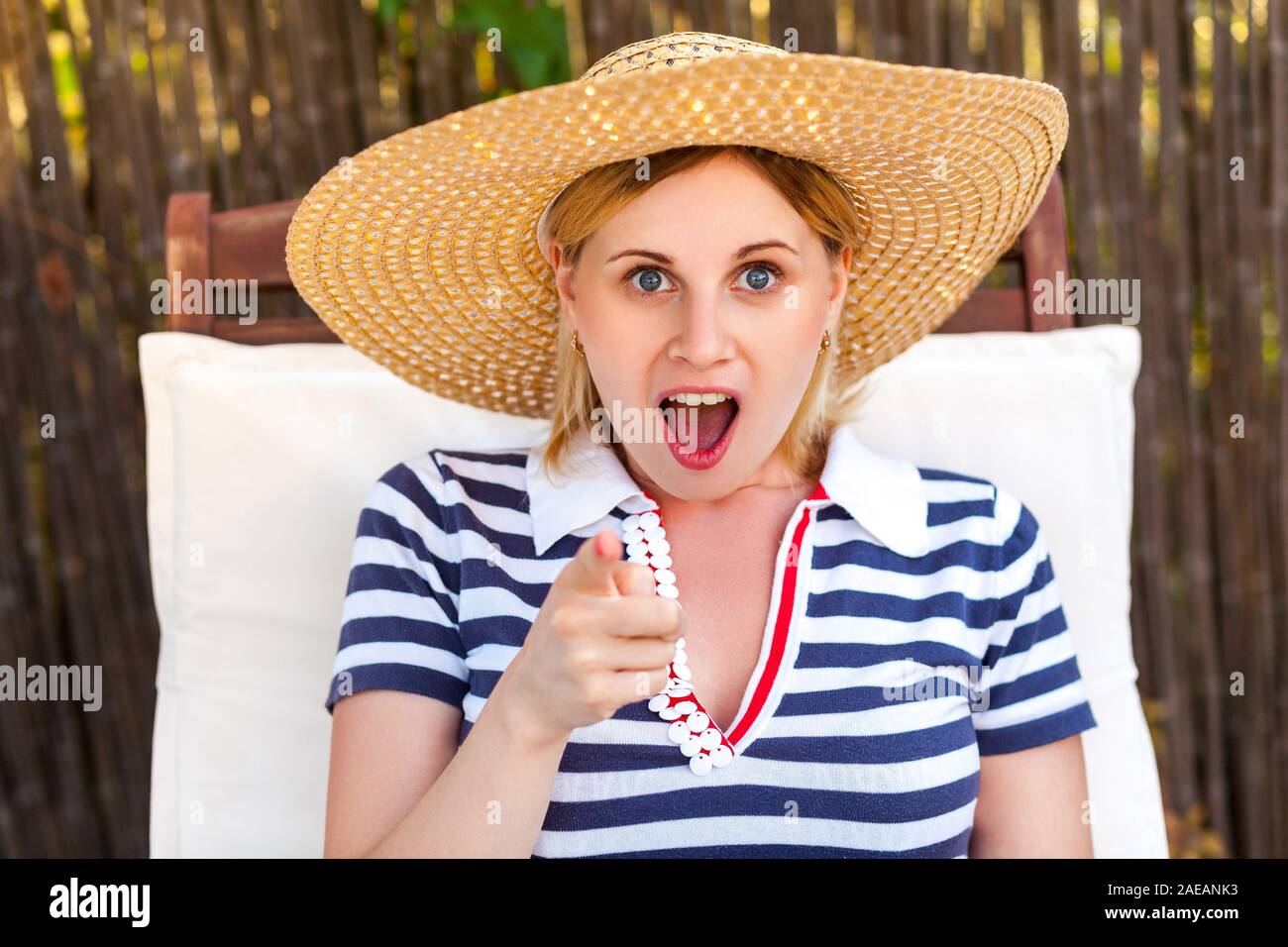 Closeup ritratto di ridere giovane donna adulta in hat e abiti seduti su confortevoli lettini da mare e puntare il dito per voi. Il concetto di stile di vita, all'aperto, estate, Foto Stock