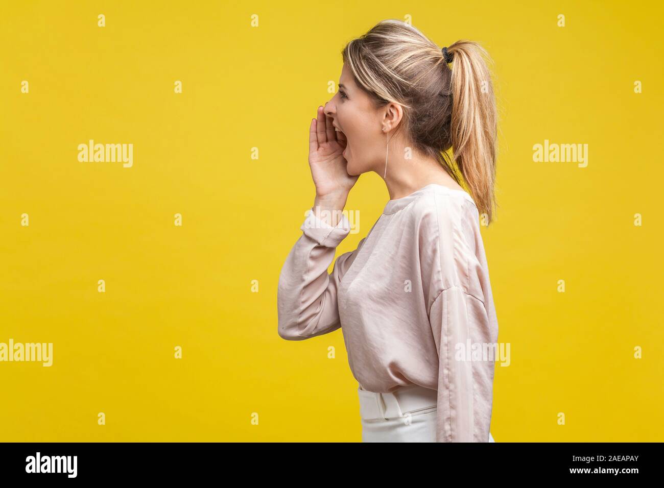 Profilo vista lato positivo della bella donna bionda con ponytale acconciatura e nel casual beige camicetta in piedi con la mano sulla bocca e gridando. in Foto Stock