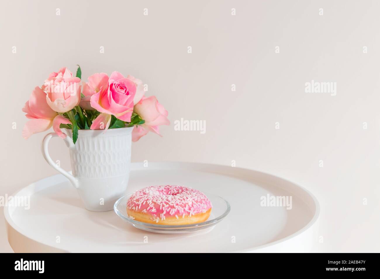 Pastel Pink roses in tazza bianca con ciambella rosa su bianco tavolino da caffè Foto Stock