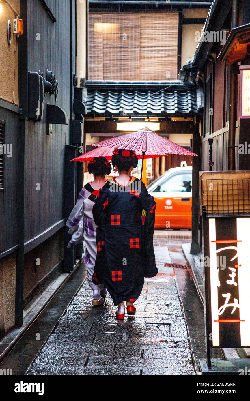 Due geisha / maiko camminando per uno stretto vicolo in possesso di una carta di riso parasol nella piovosa quartiere Gion, Kyoto, Giappone Foto Stock