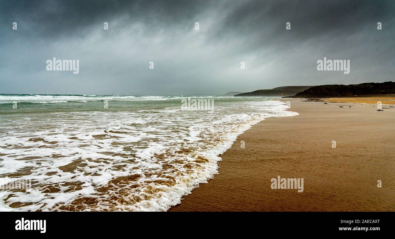 Spiaggia di sabbia in inverno in Bretagna. La schiuma bianca delle onde dell'oceano viene a morire sulla sabbia sotto un cielo scuro carico di pioggia. Foto Stock