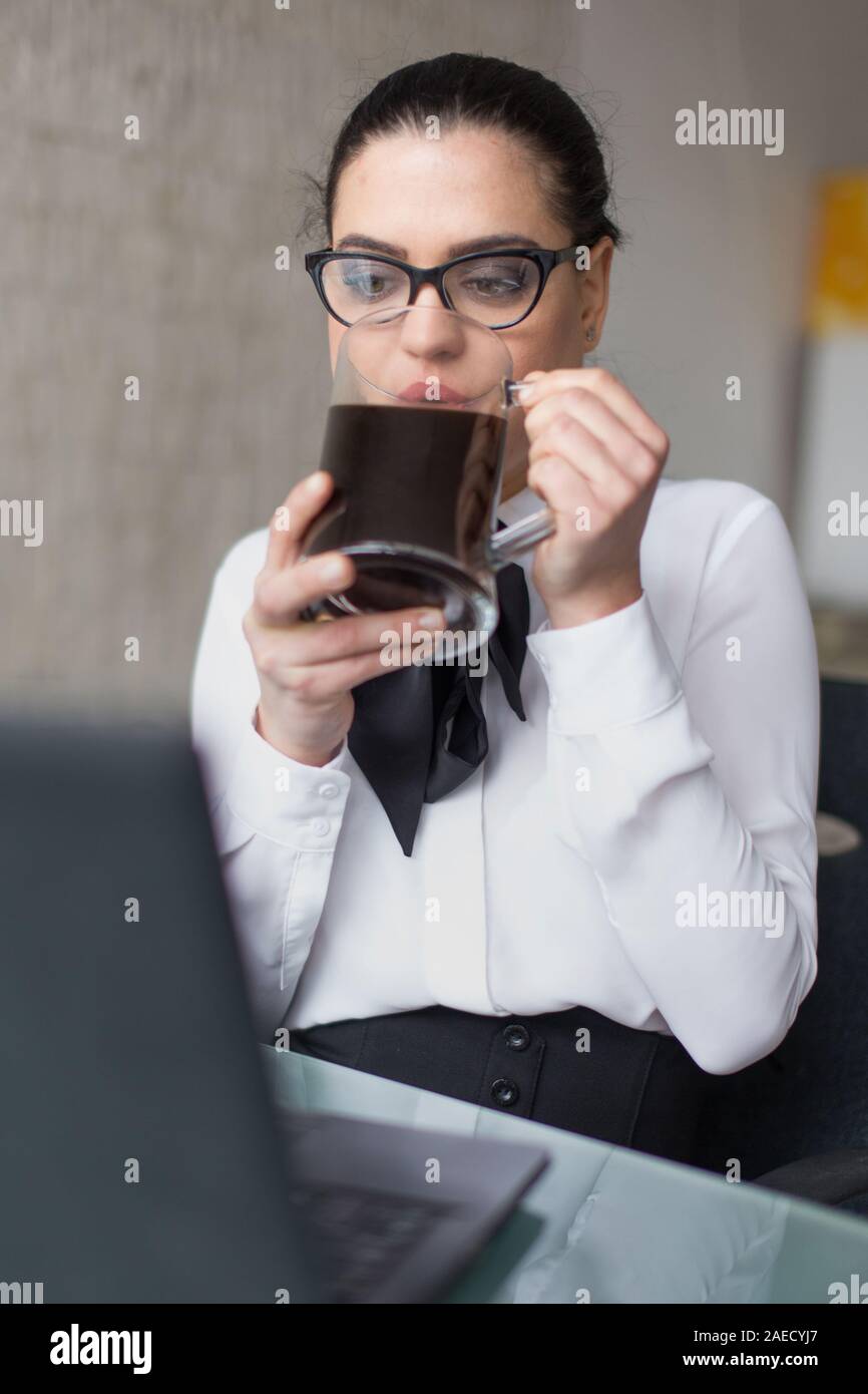 Giovane imprenditrice dipendente in abbigliamento formale di bere il caffè in ufficio, la lettura sul computer portatile Foto Stock