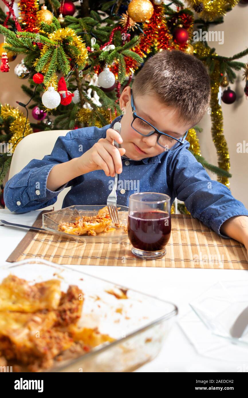 Il ragazzo con gli occhiali si siede al tavolo e il cibo di poke sulla sua forcella sulla piastra. Un bellissimo albero di Natale decorato in background. Foto Stock