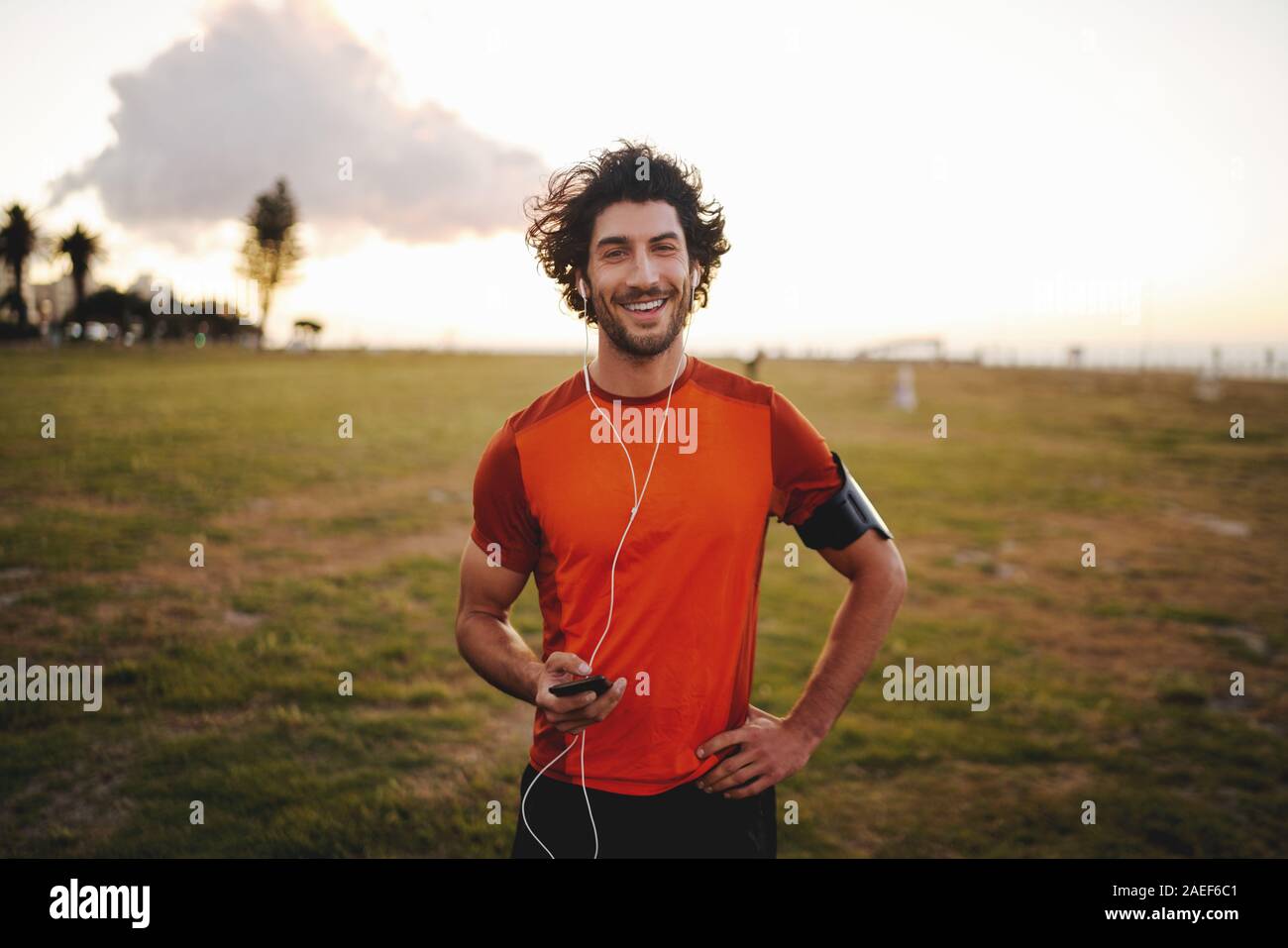 Ritratto di un felice idoneità giovane auricolari tenendo il cellulare mentre in piedi in park guardando la fotocamera Foto Stock