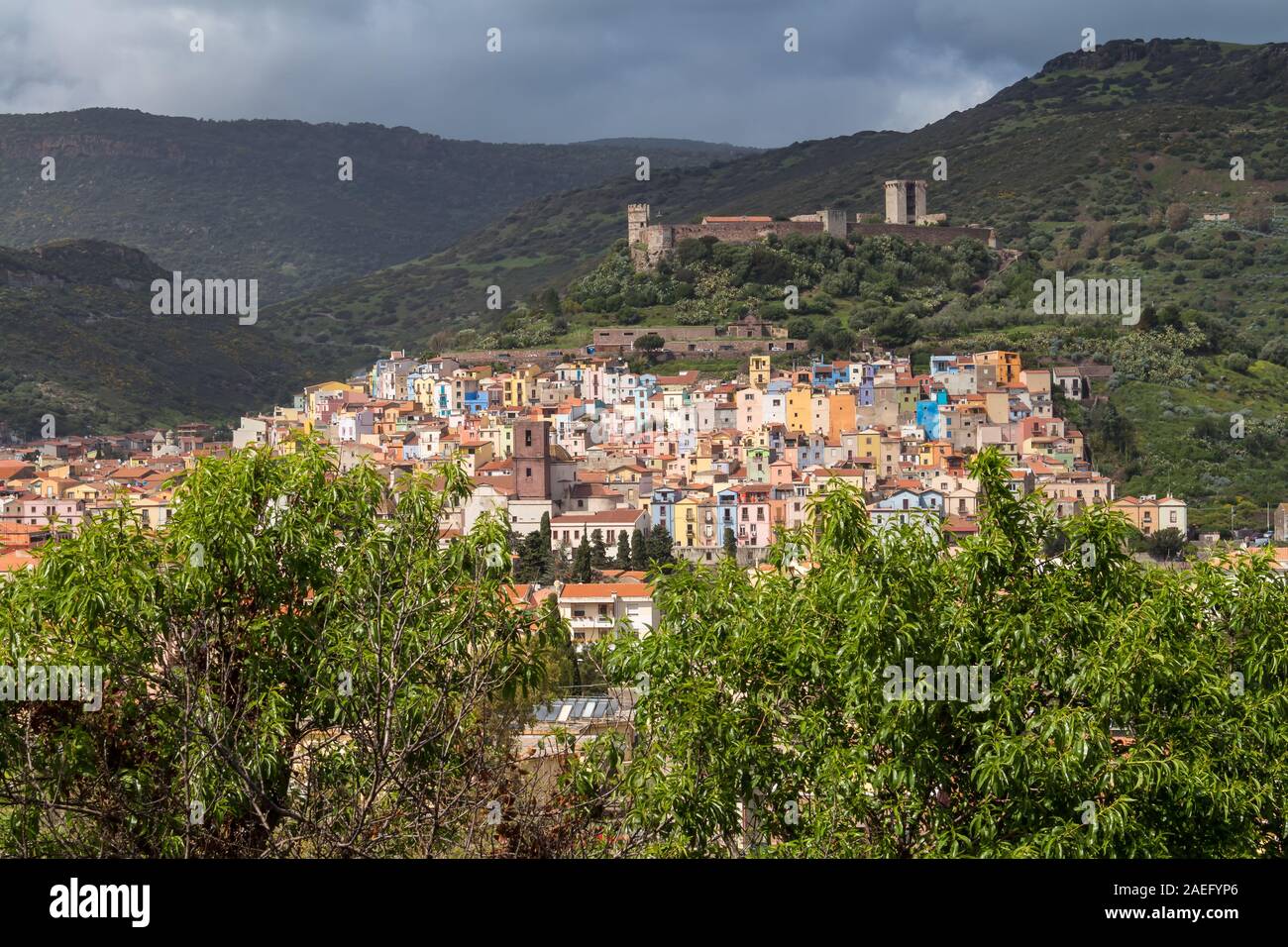 Vista aerea sul colorato piccolo città situato in una valle presso la costa del mar Mediterraneo. Fresche piante verdi, nuvoloso primavera piovosa sky. Bosa, Foto Stock