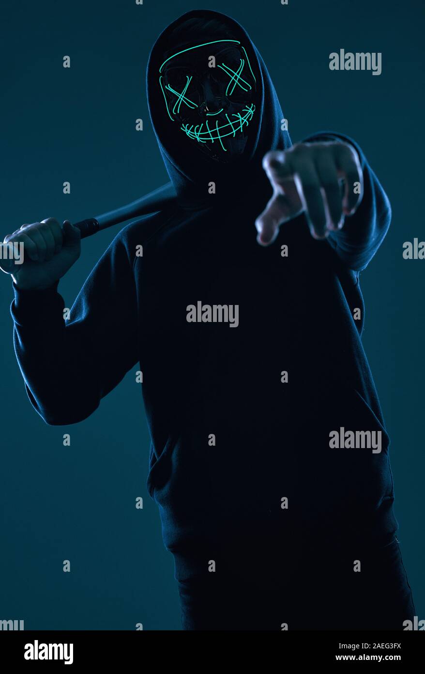 Ritratto di un anonimo uomo delinquente con la mazza da baseball in una felpa con cappuccio nero nasconde il suo volto dietro una maschera al neon. Studio shot. Foto Stock