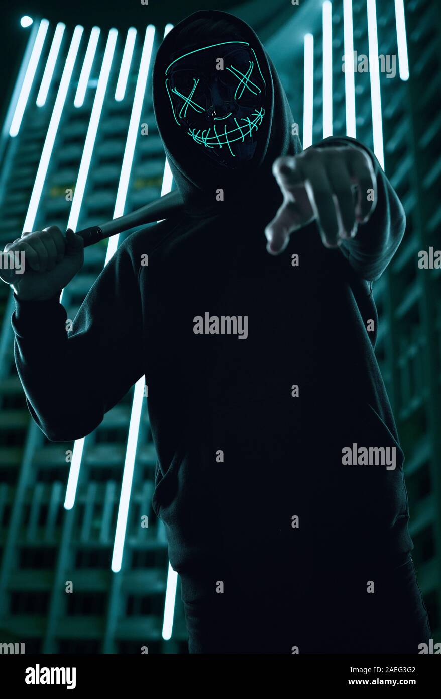 Ritratto di un anonimo uomo delinquente con la mazza da baseball in una felpa con cappuccio nero nasconde il suo volto dietro una maschera al neon. Città luminosa sullo sfondo Foto Stock