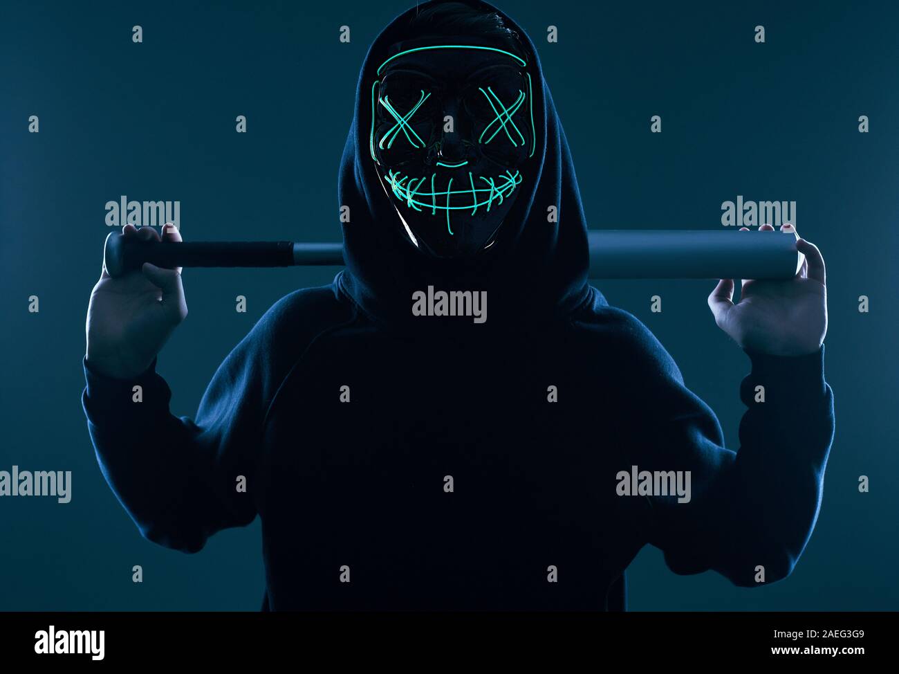 Ritratto di un anonimo uomo delinquente con la mazza da baseball in una felpa con cappuccio nero nasconde il suo volto dietro una maschera al neon. Studio shot. Foto Stock