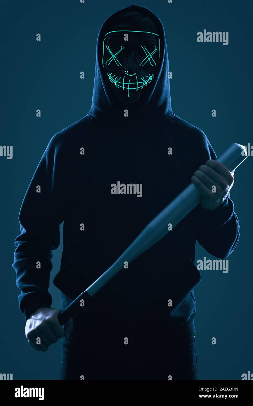 Ritratto di un anonimo uomo delinquente con la mazza da baseball in una felpa con cappuccio nero nasconde il suo volto dietro una maschera al neon. Studio shot. Foto Stock