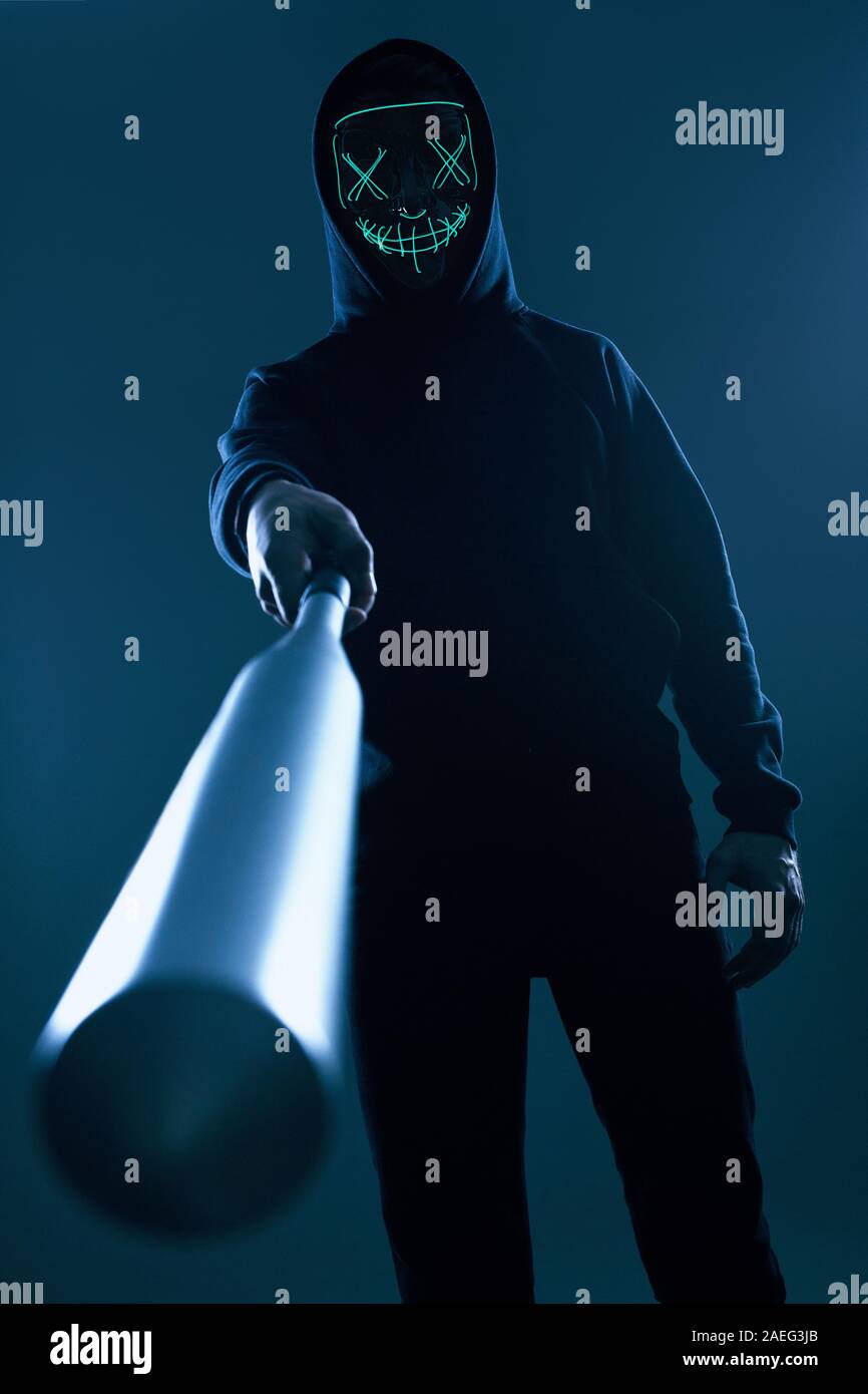 Ritratto di un anonimo uomo delinquente con la mazza da baseball in una felpa con cappuccio nero nasconde il suo volto dietro una maschera al neon. Angolo inferiore Foto Stock