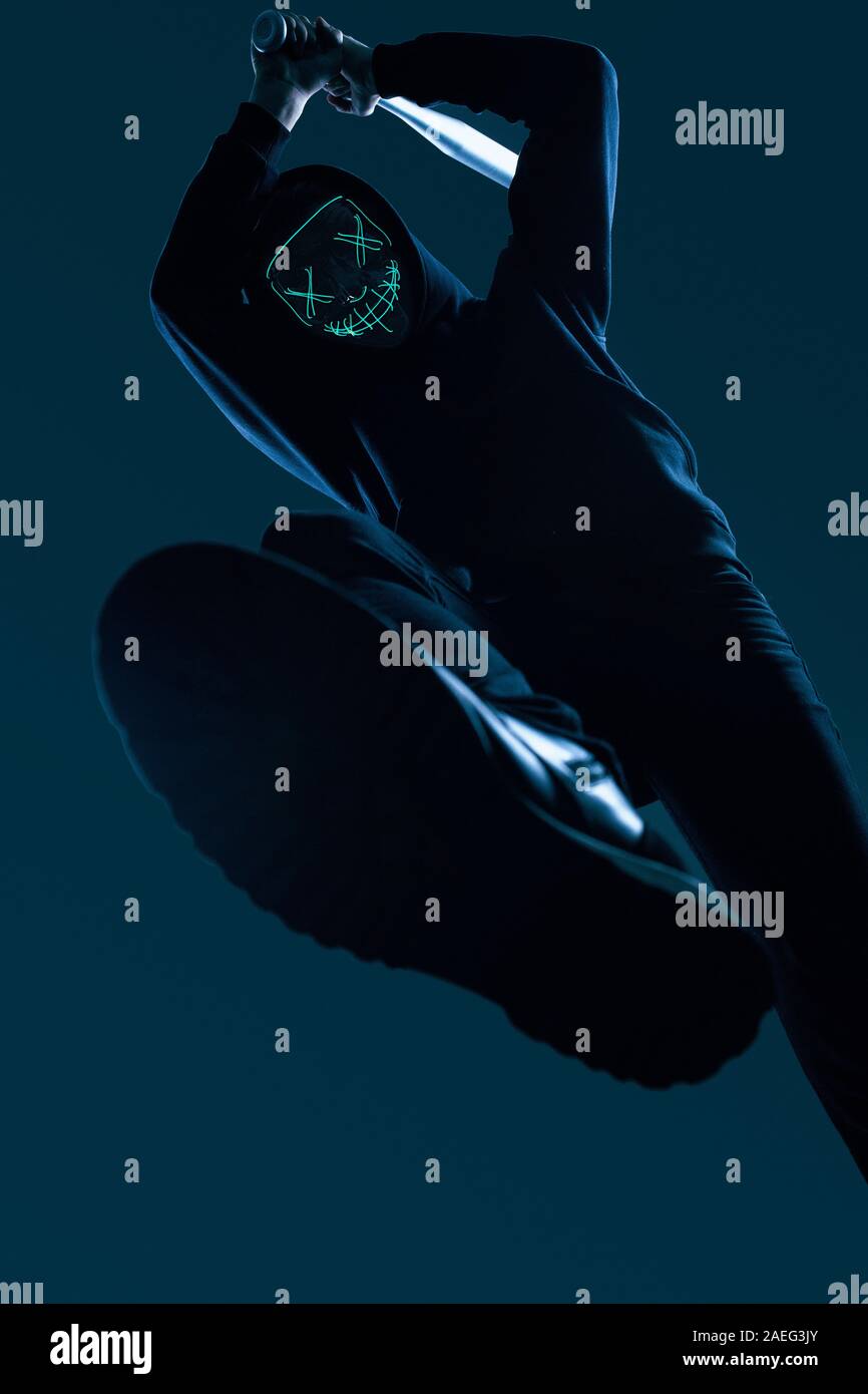 Ritratto di un anonimo uomo delinquente con la mazza da baseball in una felpa con cappuccio nero nasconde il suo volto dietro una maschera al neon. Angolo inferiore Foto Stock