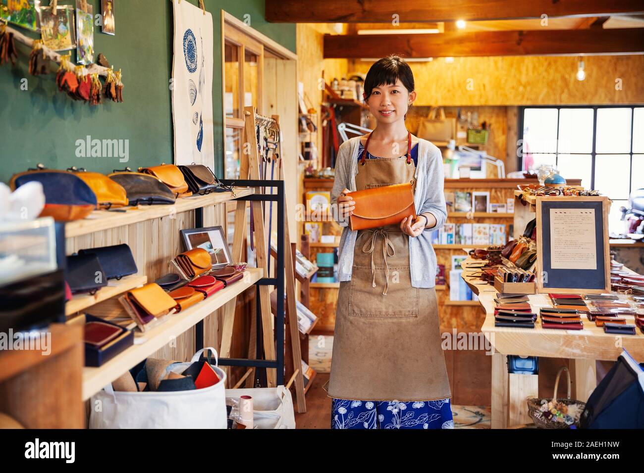 Donna Giapponese che indossa il grembiule in piedi in un negozio di cuoio, tenendo in pelle marrone pochette, sorridente alla fotocamera. Foto Stock