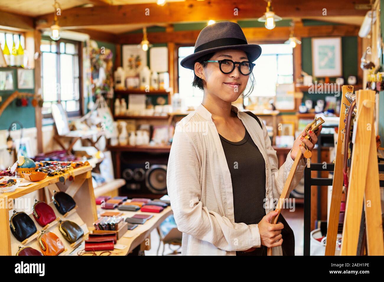 Donna Giapponese che indossa un cappello e occhiali in piedi in un negozio di cuoio, tenendo cintura in cuoio. Foto Stock