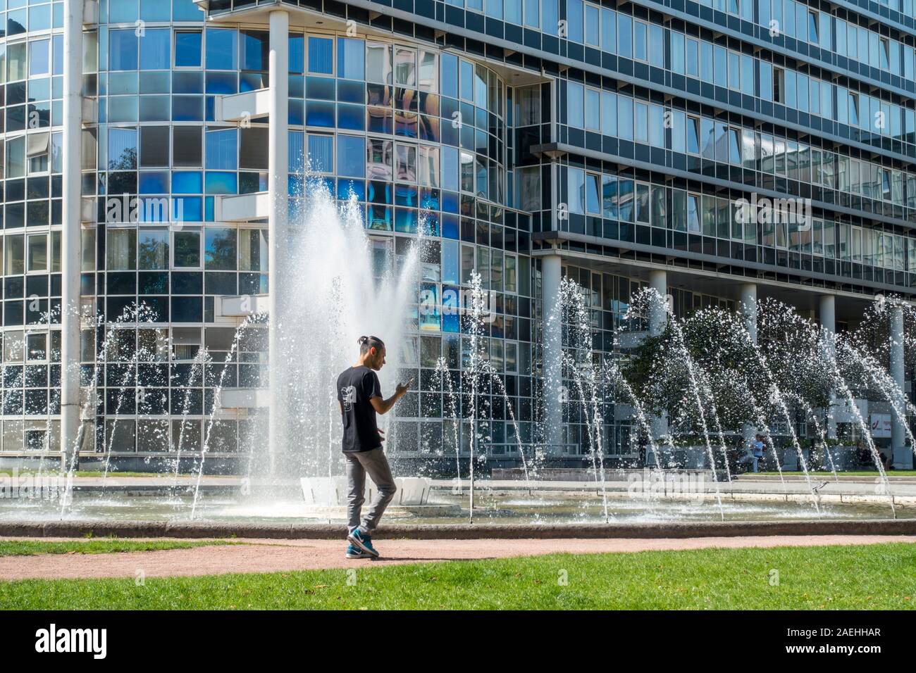 Uomo che cammina durante la lettura di smartphone nella parte anteriore della fontana nel re Albert Park, il parco della città nel distretto del Sud a Gand, Fiandre Orientali, Belgio Foto Stock