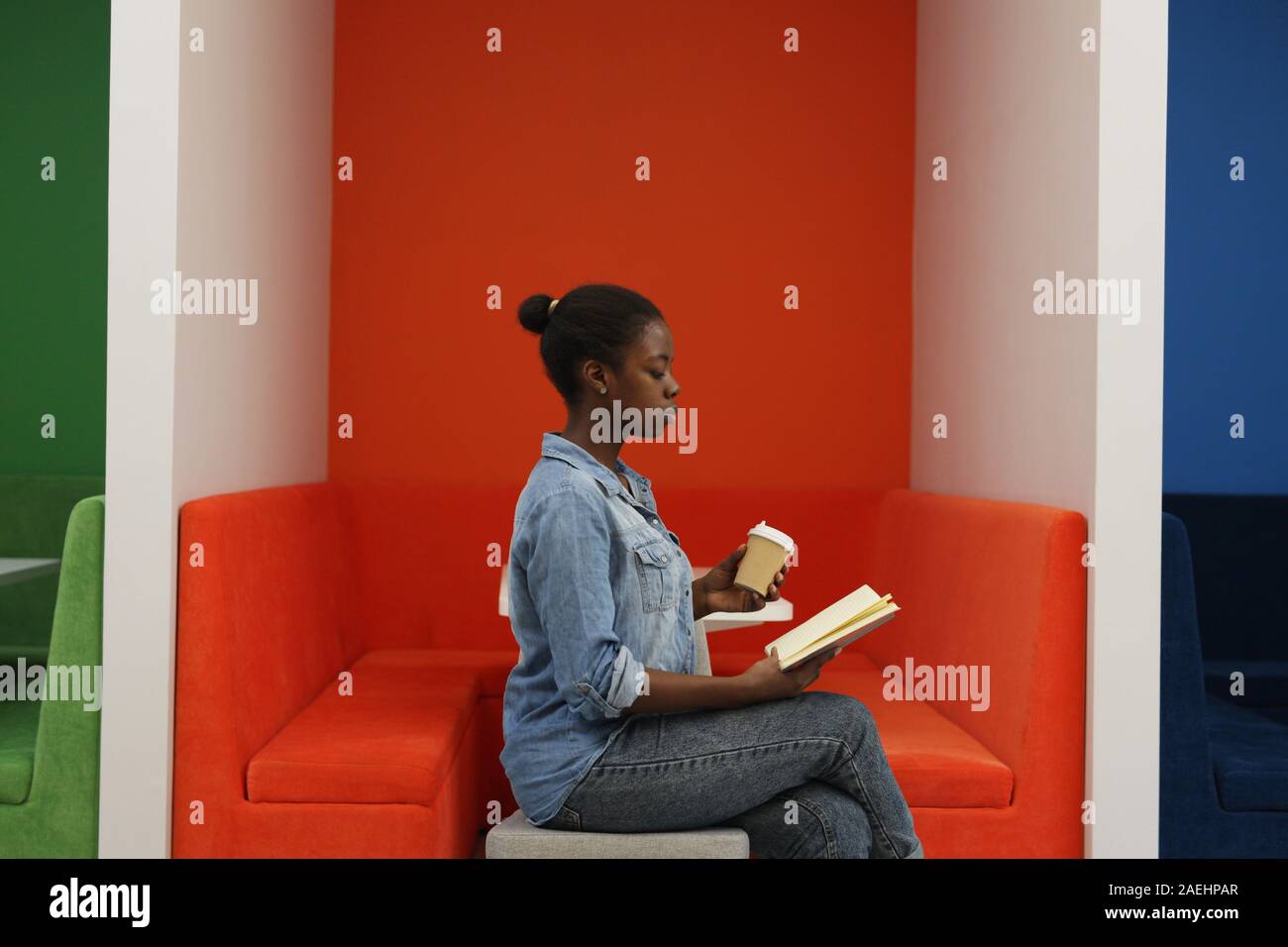 Vista laterale dello studente africano in jeans seduta di usura ed a concentrarsi sulla lettura di un libro e tenendo una tazza di caffè in mano Foto Stock