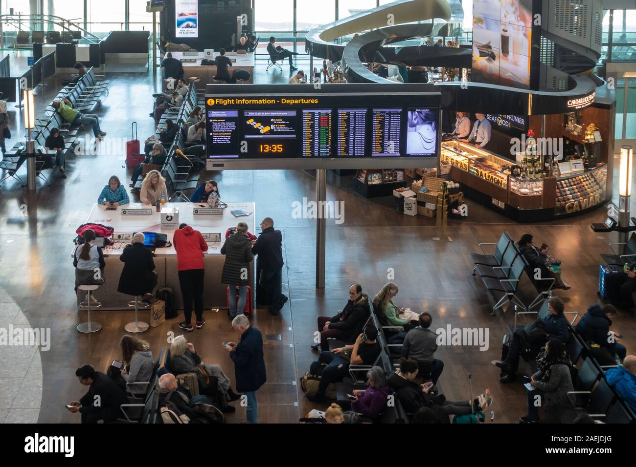 Un volo information board nel mezzo di panche pieno di persone sedute, attesa per i loro voli a Londra Heathrow Airport, terminale 5. Foto Stock