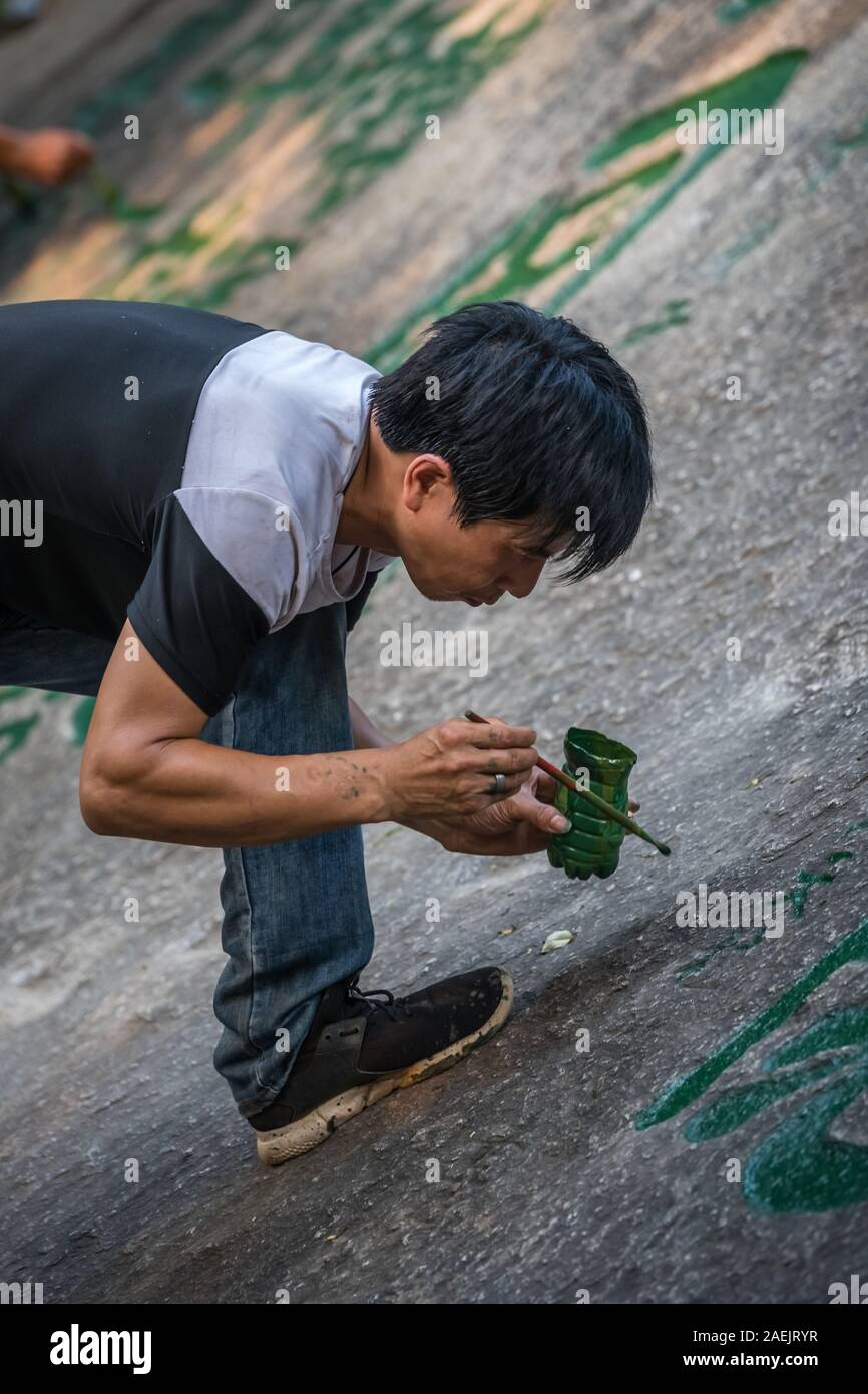 Huashan, Cina - Agosto 2019 : uomo pittura calligrafia cinese caratteri su un grande masso nel cortile di un tempio buddista ai piedi di Hu Foto Stock