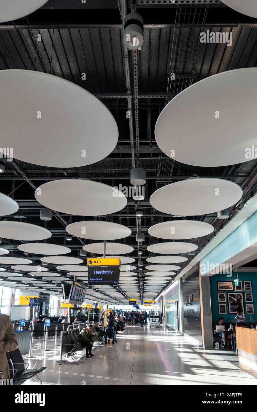 La gente in attesa all'aeroporto gates al Terminal 5 di Londra Heathrow. I dischi decorativi decorare il soffitto. Foto Stock