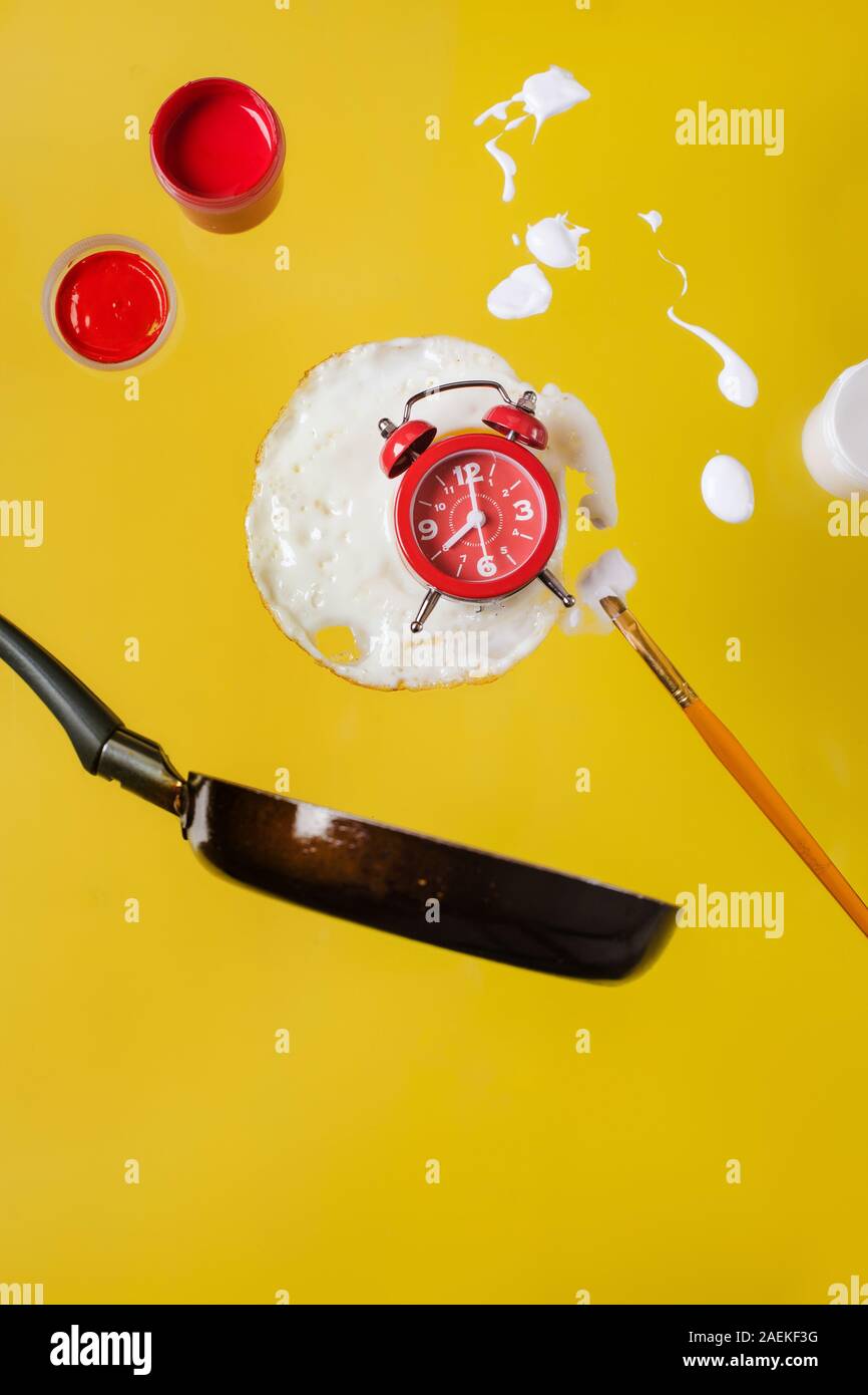 Uovo fritto e la padella con un orologio rosso, barattoli di vernice e vernici a spruzzo e a pennello levitare in aria. Foto Stock