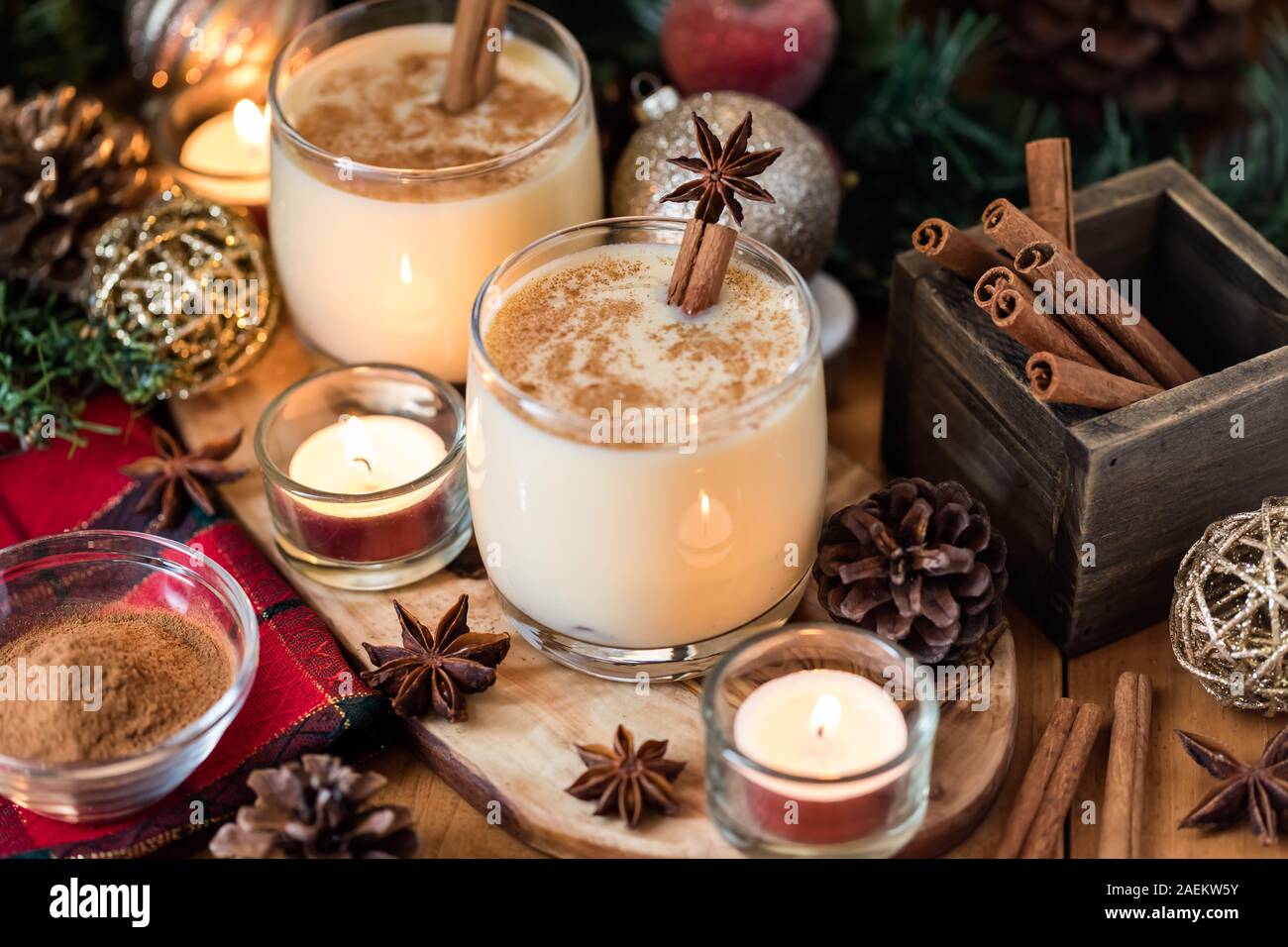 Una vista ravvicinata di due bicchieri di uovo nog con cannella e anice stellato e circondato da decorazioni natalizie e candele. Foto Stock
