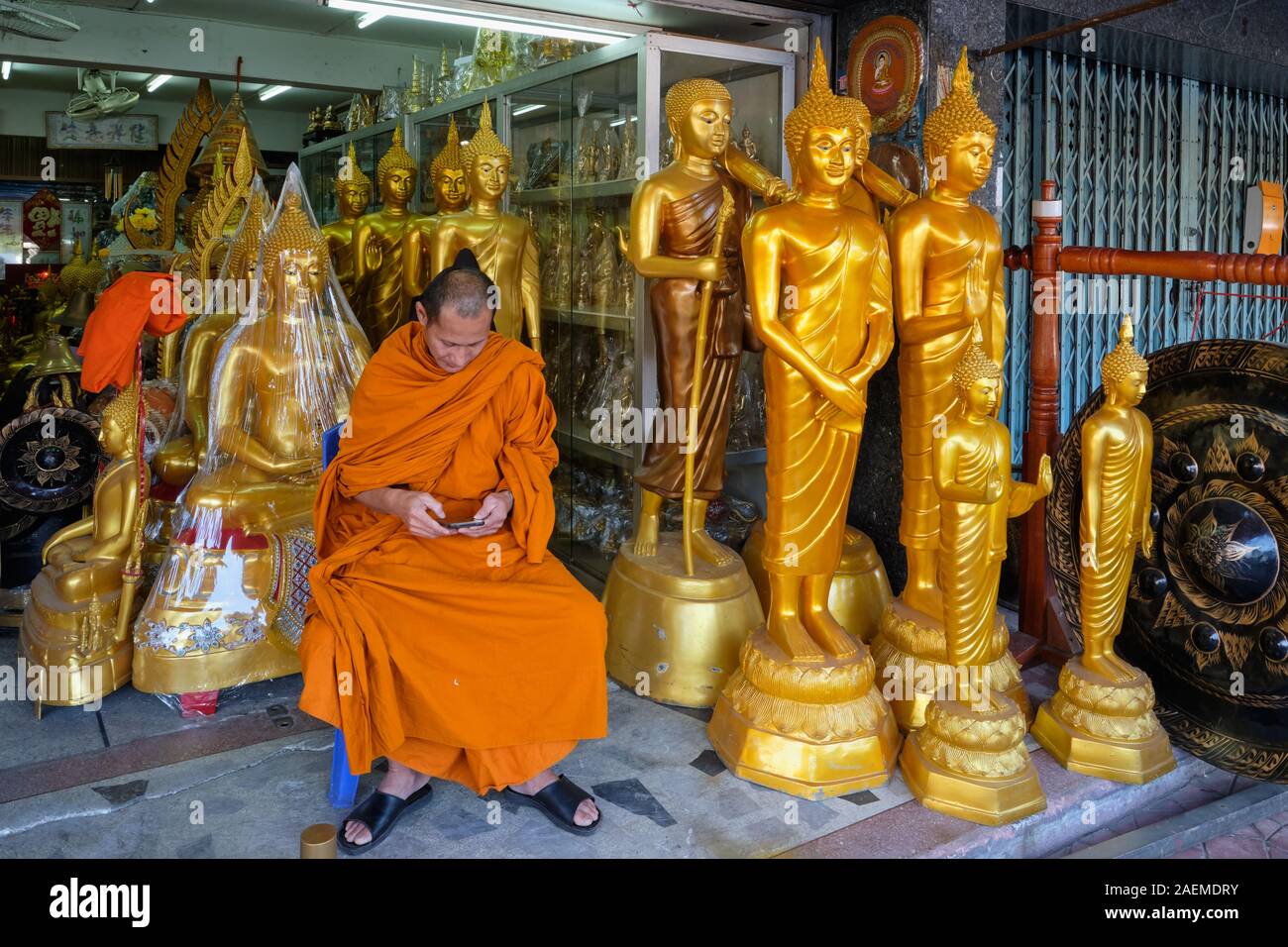 Un monaco buddista si siede tra statue di Buddha in un negozio per gli artefatti buddista, intensamente il peering presso il suo mobile; Bamrung Muang Rd., Bangkok, Thailandia Foto Stock