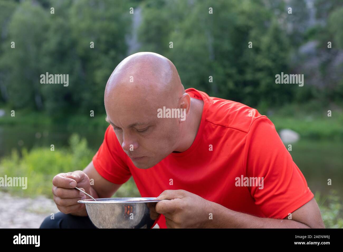 Uomo calvo, soldato in una t-shirt rossa mangia da una ciotola di stagno per esterno contro un paesaggio forestale Foto Stock