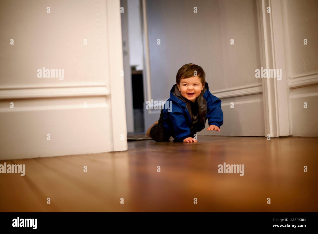 Baby boy sorrisi come egli esegue la scansione attraverso una porta su un pavimento di legno. Foto Stock