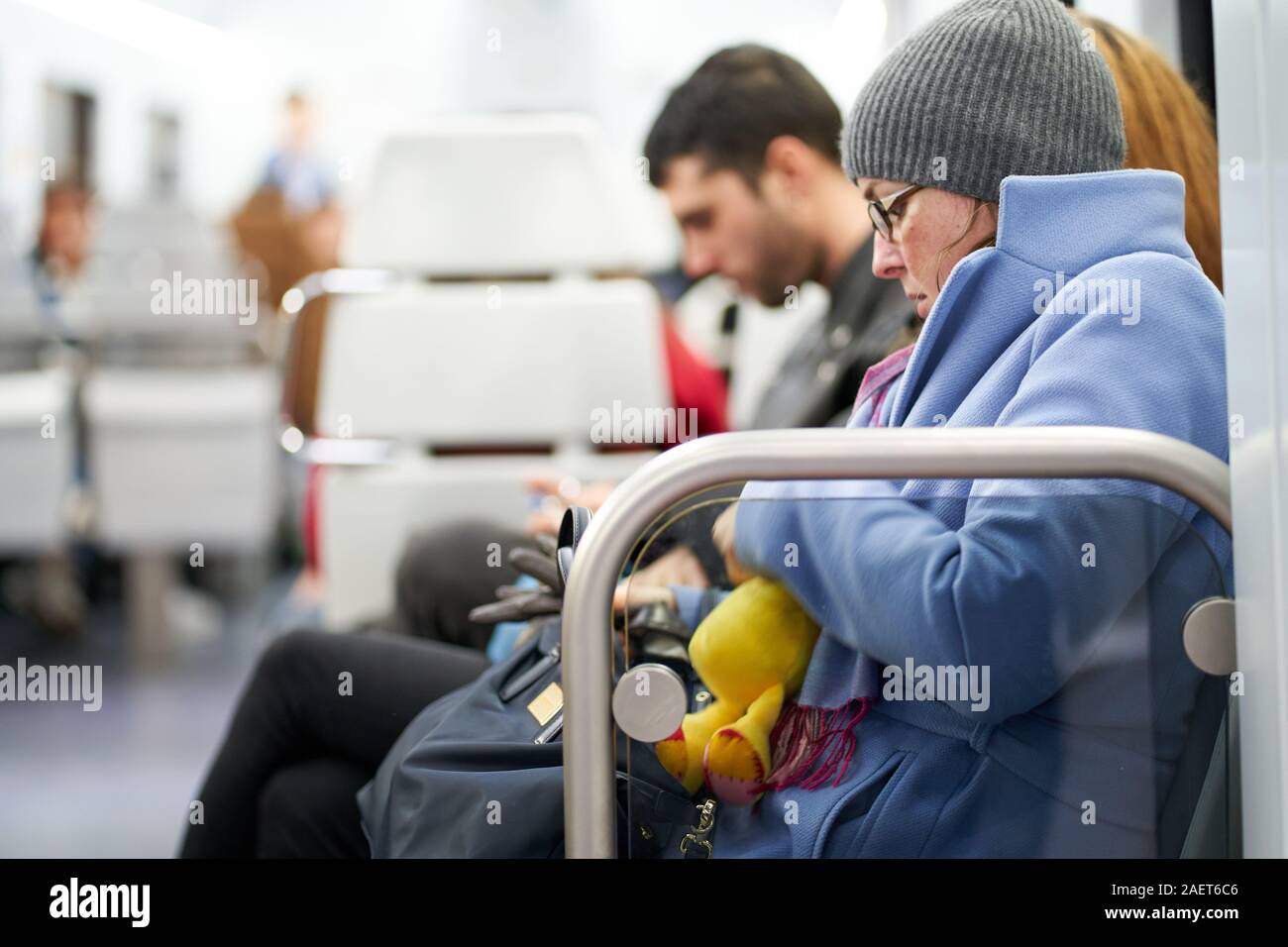 Una giovane ragazza seduta su un treno in un mantello blu con un beanie cappello e occhiali assiste alla sua borsa mentre tiene un anatra giallo Foto Stock
